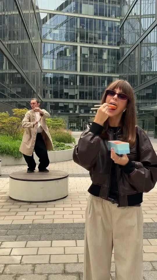 Sergei eating in front of a glass building while Elvira dances behind him